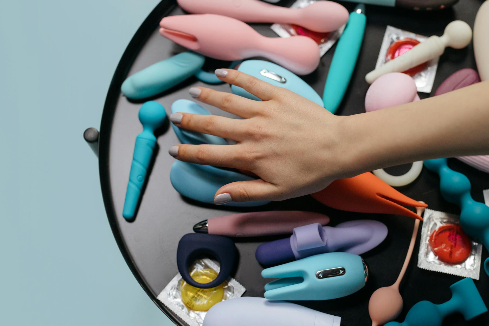 A hand reaching over a variety of colorful sex toys and vibrators arranged on a table.